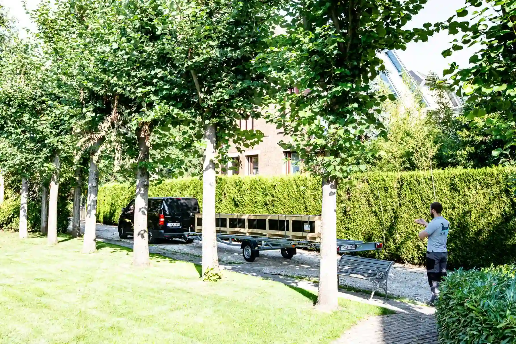 linstallation-chez-juliette-leon-1 Un véhicule noir avec une remorque est stationné sur une allée bordée d'arbres, près d'une haie verte et d'un banc en métal.
