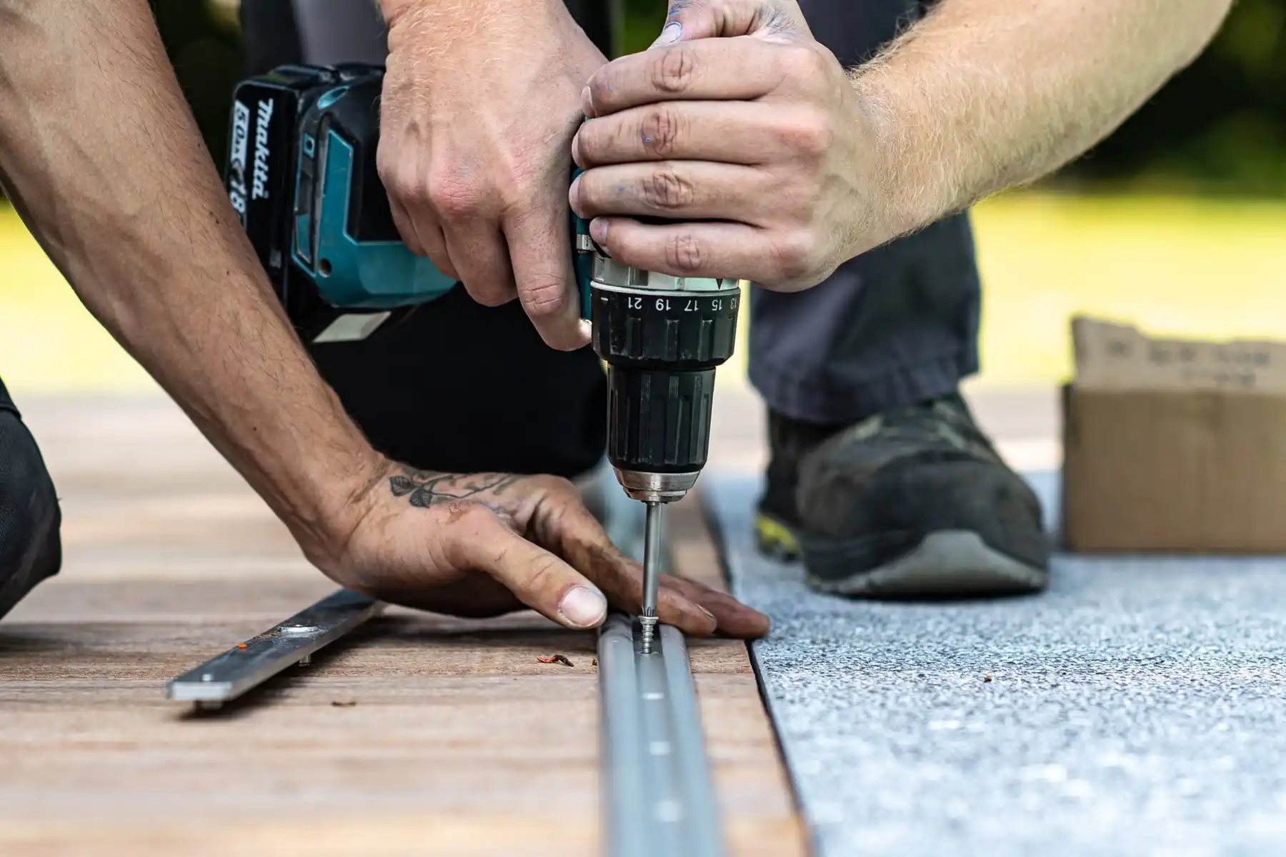 Deux mains installent une vis avec une perceuse sans fil sur un rail métallique posé sur une surface en bois et tapis gris.