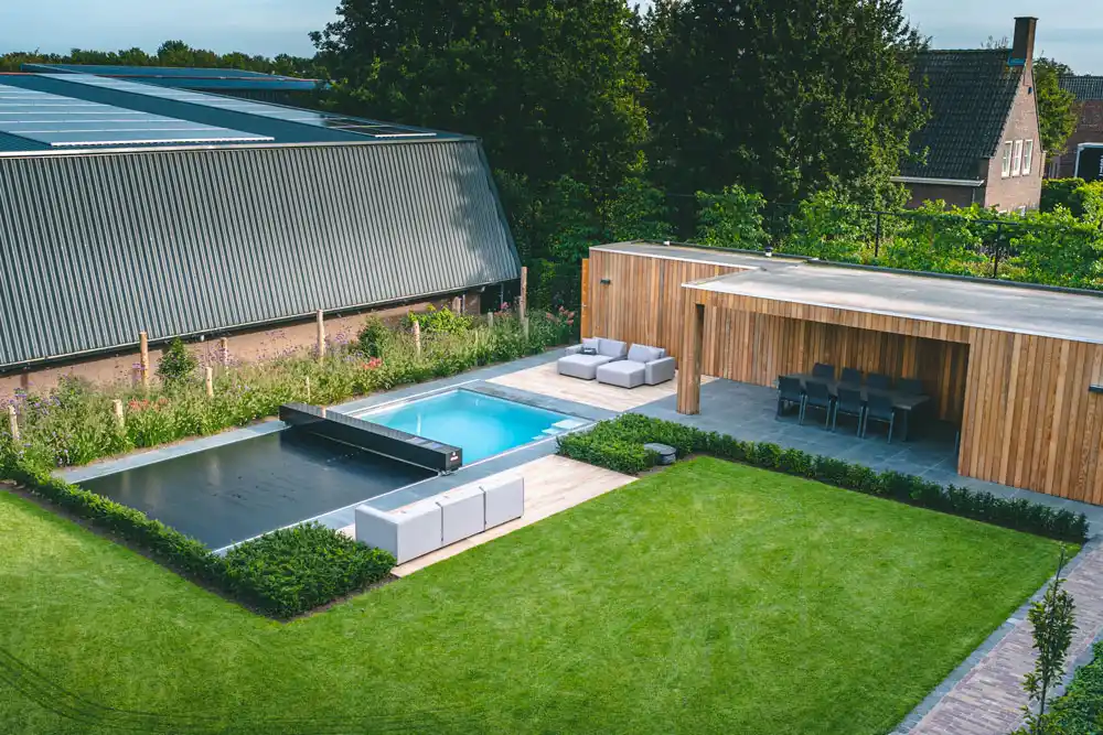 veerle-et-jan-coverseal Une piscine avec couverture grise est entourée de mobilier de jardin et d'une terrasse en bois, avec pelouse verte et haies soignées.
