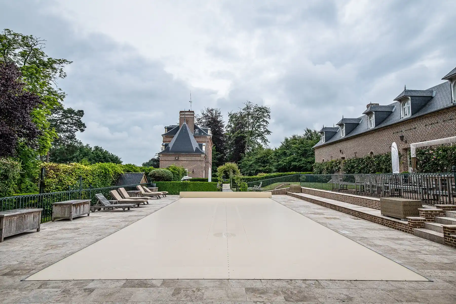 Chez-Camille-et-Arthur-2 Piscine couverte de beige entourée de chaises longues, située devant un bâtiment en briques avec un ciel nuageux.