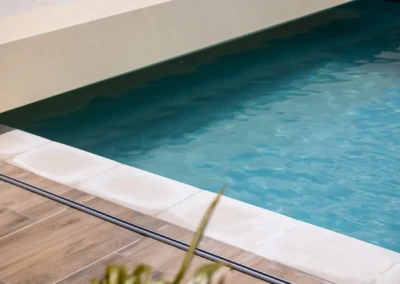 Une piscine aux eaux bleues est bordée de dalles blanches et d'un sol en bois clair.
