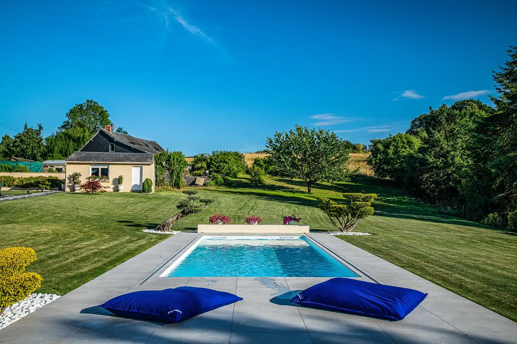 Chez-Gwendoline-et-François-3 Maison beige avec toit noir, piscine entourée de coussins bleus, pelouse verte et arbres en arrière-plan sous ciel bleu.