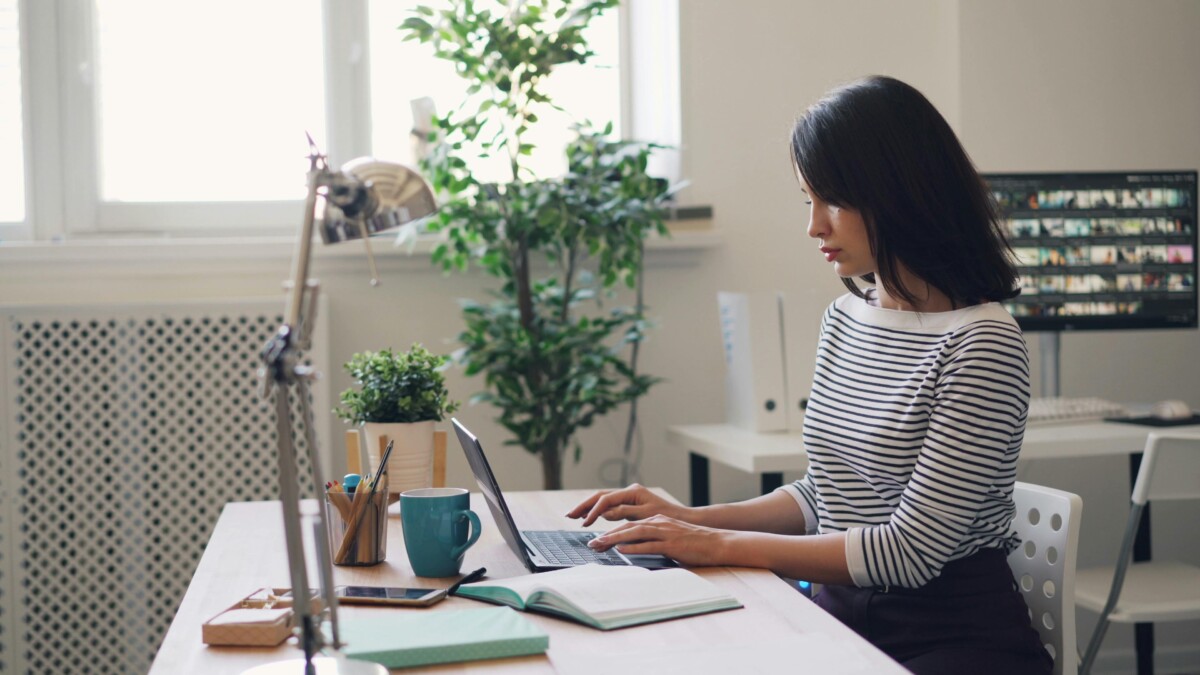 Une personne travaille sur un ordinateur portable à un bureau, avec une tasse bleue et des plantes en arrière-plan.