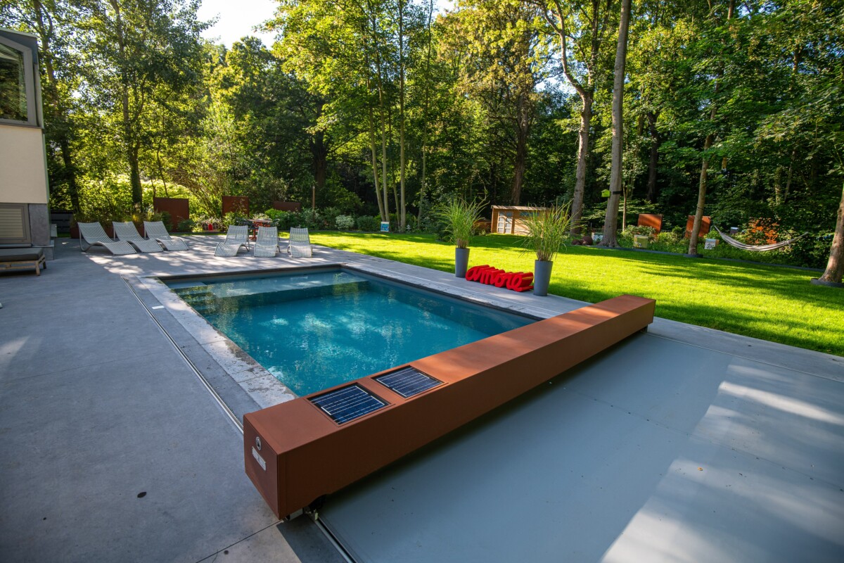Corten (18)-min Piscine rectangulaire entourée de chaises longues blanches, bordée par une terrasse en béton et une pelouse verte avec des arbres en arrière-plan.