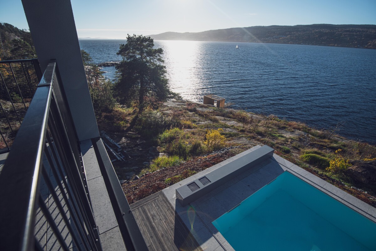 DROBAK – SUN (8) Vue d'un balcon surplombant une piscine bleue bordée de dalles grises, avec un paysage côtier et un arbre en arrière-plan.