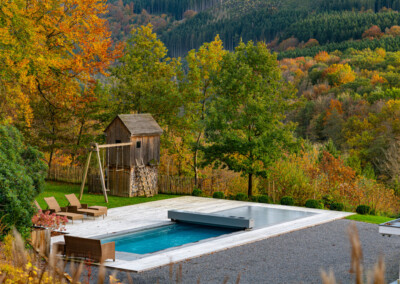 Une piscine rectangulaire bordée de chaises longues brunes, entourée d'arbres aux couleurs automnales et d'une cabane en bois.