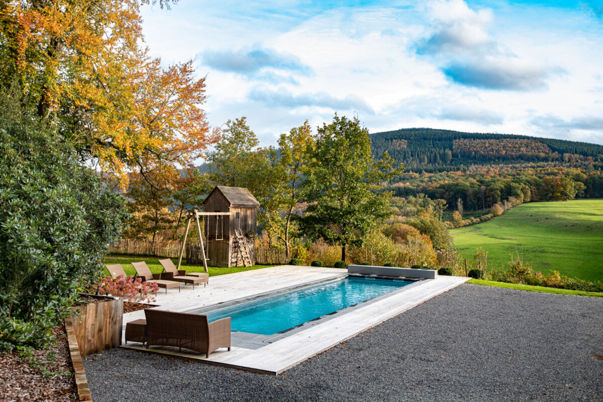LANDSCAPE_-_FALL_(8)-_réal Une piscine rectangulaire entourée de chaises longues marron, avec un abri en bois et des arbres aux couleurs automnales en arrière-plan.