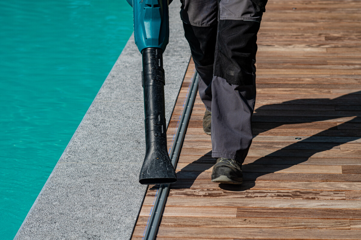PIERRE-ET-BOIS (37) Un homme en pantalon gris utilise un souffleur noir sur une terrasse en bois près d'une piscine turquoise.