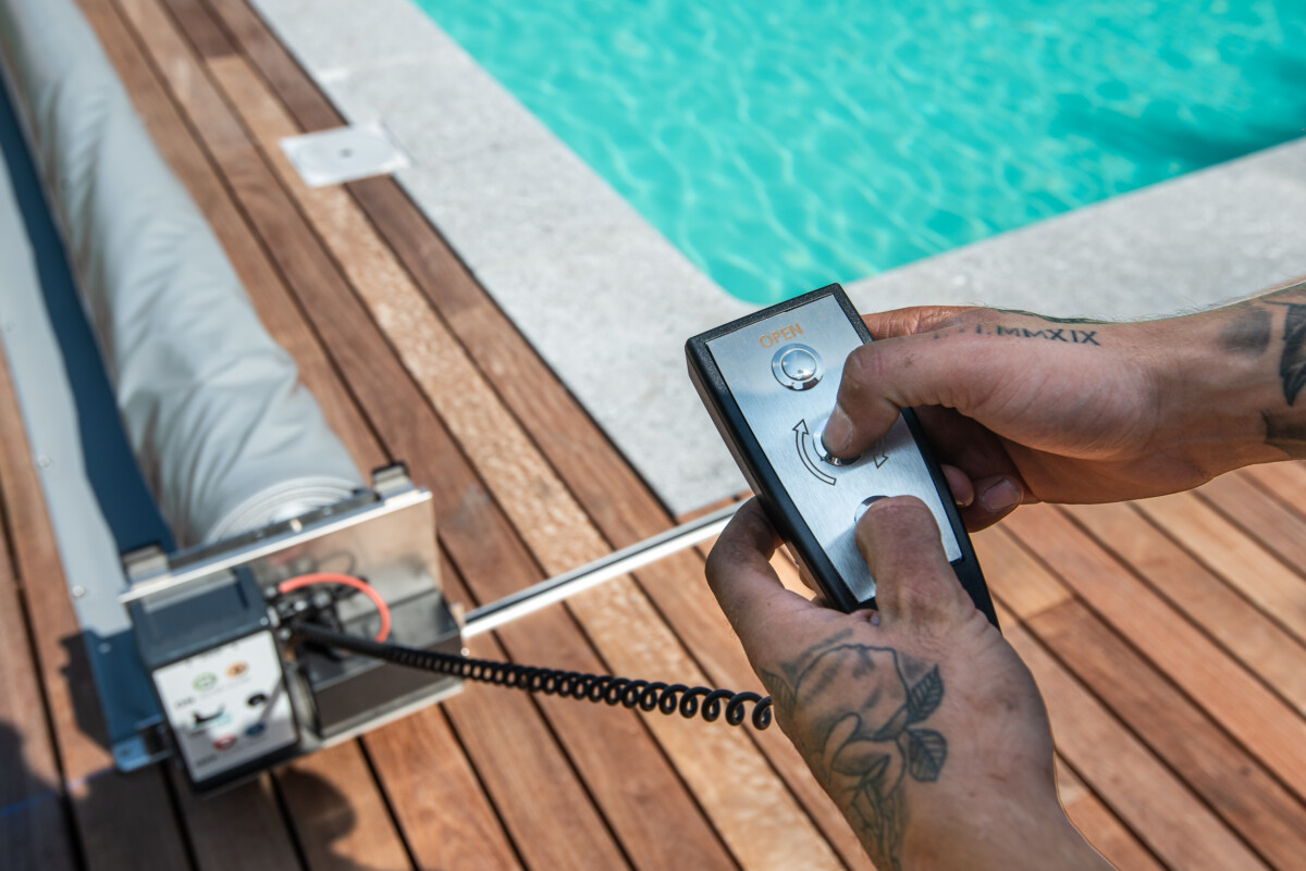 PIERRE-ET-BOIS (61) Une télécommande noire est utilisée pour ouvrir une couverture de piscine grise, située sur une terrasse en bois près de l'eau turquoise.