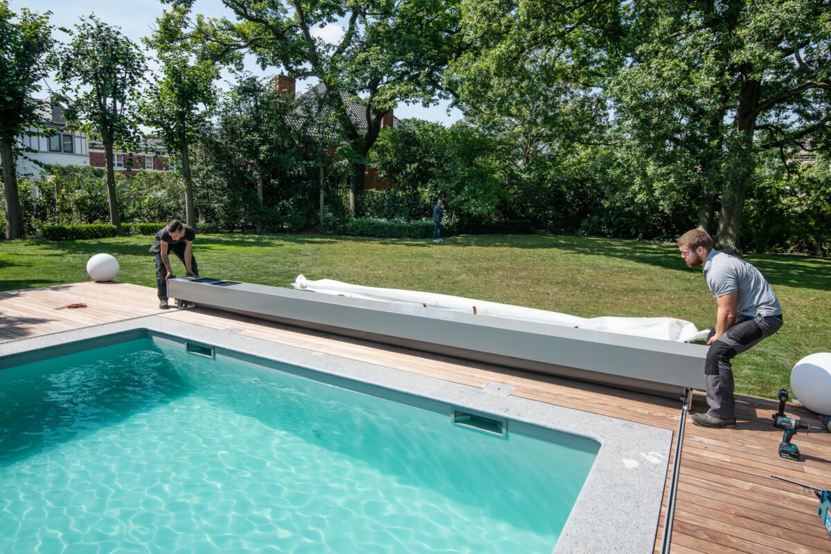 PIERRE-ET-BOIS (64) Deux personnes installent un volet roulant blanc sur une piscine entourée de bois, avec un jardin verdoyant en arrière-plan.