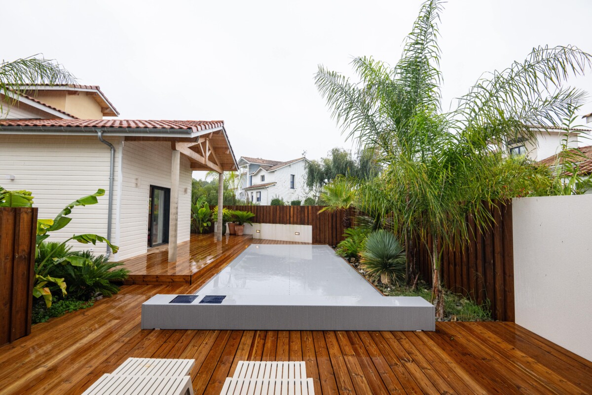 Terrasse en bois avec piscine couverte, bordée de palmiers et d'une maison blanche au toit en tuiles rouges.