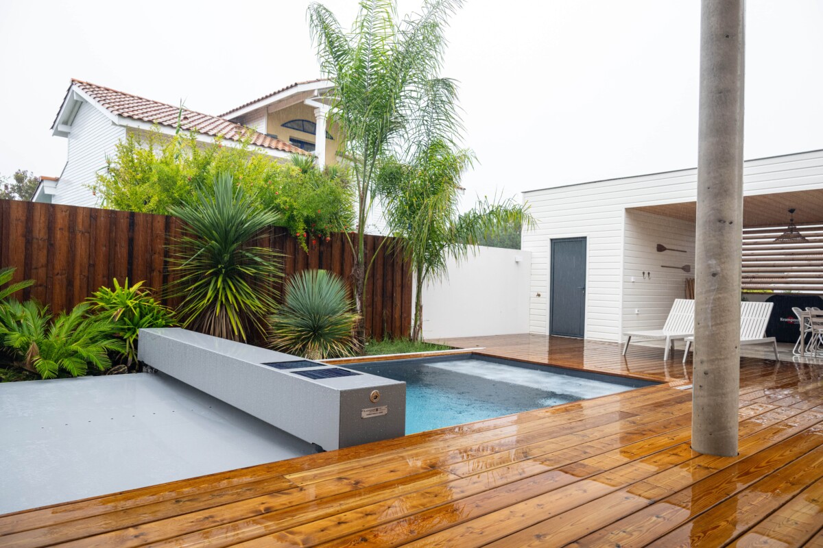 Tropical_rain31-min Une piscine entourée de palmiers et de plantes, avec une terrasse en bois mouillée et un bâtiment blanc en arrière-plan.