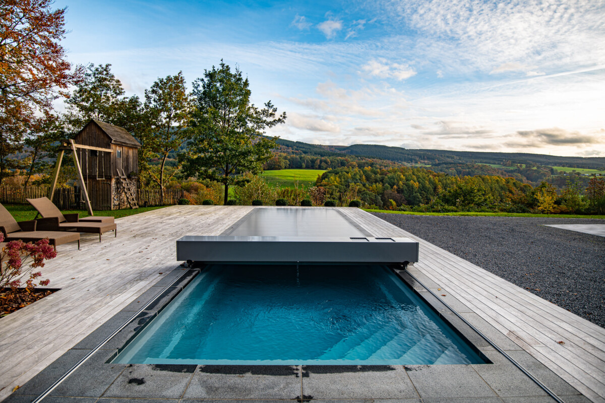 VUE-AUTOMNE_(6)-_Réal Piscine couverte par une structure grise sur une terrasse en bois, avec des arbres et des collines en arrière-plan sous un ciel partiellement nuageux.