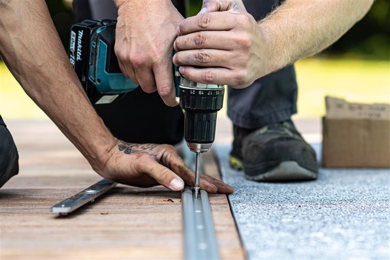 WOOD-AND-STONE (18) (1) Deux mains installent une vis avec une perceuse sur un rail métallique posé sur du bois, avec un carton à proximité.